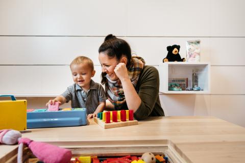 A small child smiles as he plays with toys at a table with a female caregiver or family member watching nearby.