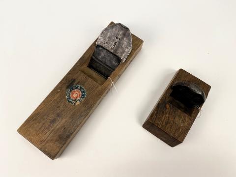 Two wooden plane tools sit on a white talbe. One has a round sticker. Both have Japanese characters on the blades. 