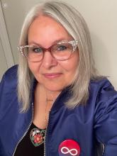  Connie Kulhavy looking out of the frame with a small smile on her face. She has mid-length white hair, pink cat-eye classes and a blue jacket.