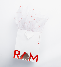 A white gift bag with white tissue paper coming out of the top. The bag has the letters RAM in red with a symbol of a mammoth walking through the A.